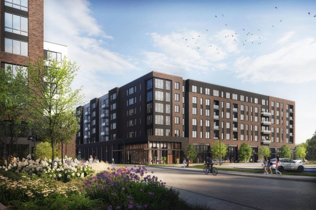 A modern mid-rise apartment building with large windows rises near Rambler Riverfront, surrounded by landscaped gardens, trees, and people walking or biking on a sunny day. Birds fly in the blue sky above.