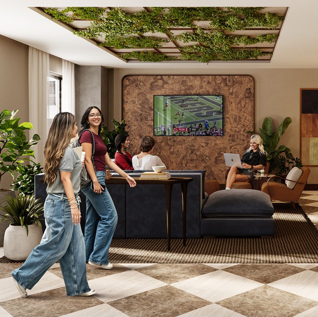 A modern lounge with lush plants, where five women relax—two walk and smile in the foreground, while three sit on sofas at Purdue off campus housing; one uses a laptop, and a TV displays a football game on the wall behind them.