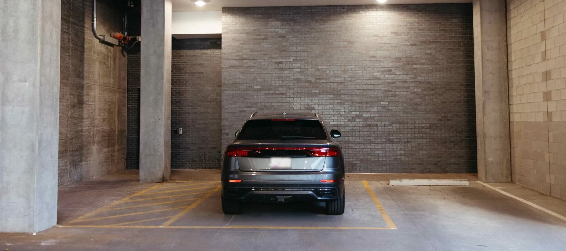 A gray SUV is parked in a mostly empty indoor parking garage, centered between yellow lines in front of a brick wall—an ideal feature for Purdue off campus housing, with overhead lights and sturdy concrete walls providing security.