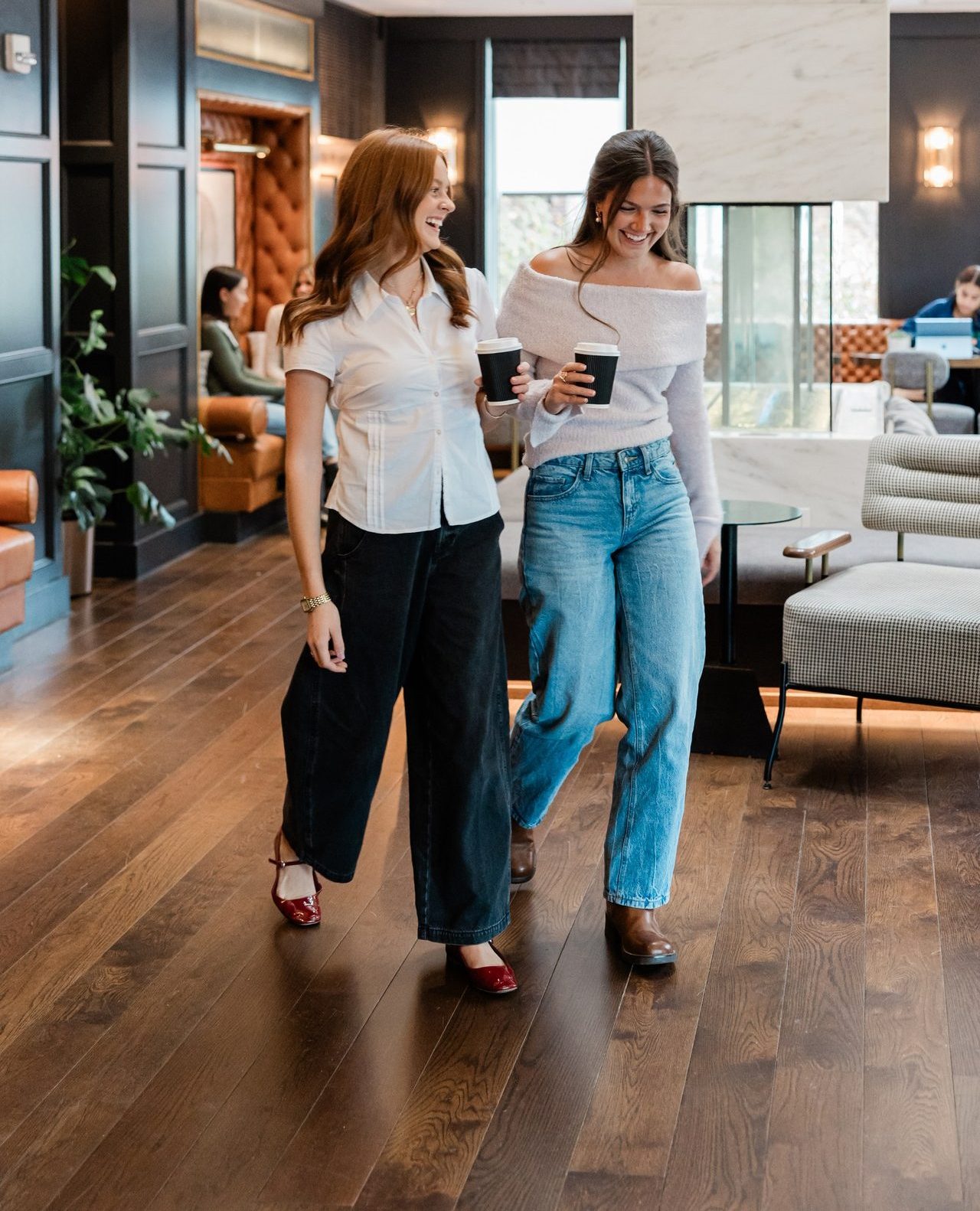 Two women walk together indoors, smiling and holding coffee cups. One wears a white blouse and black wide-leg pants; the other wears an off-shoulder top and blue jeans. The setting is a modern, stylish lounge with wood floors and furniture.