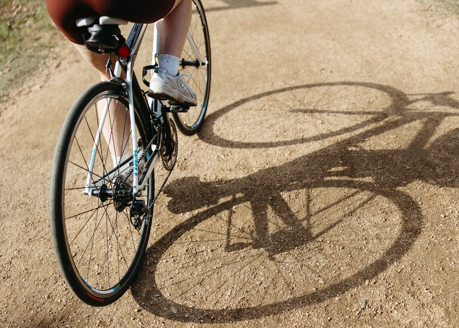 Close-up of a person riding a bicycle on a dirt path near Purdue off campus housing, showing the bike’s rear wheel and the rider’s legs. The sun casts a clear shadow of the bicycle and rider on the ground.