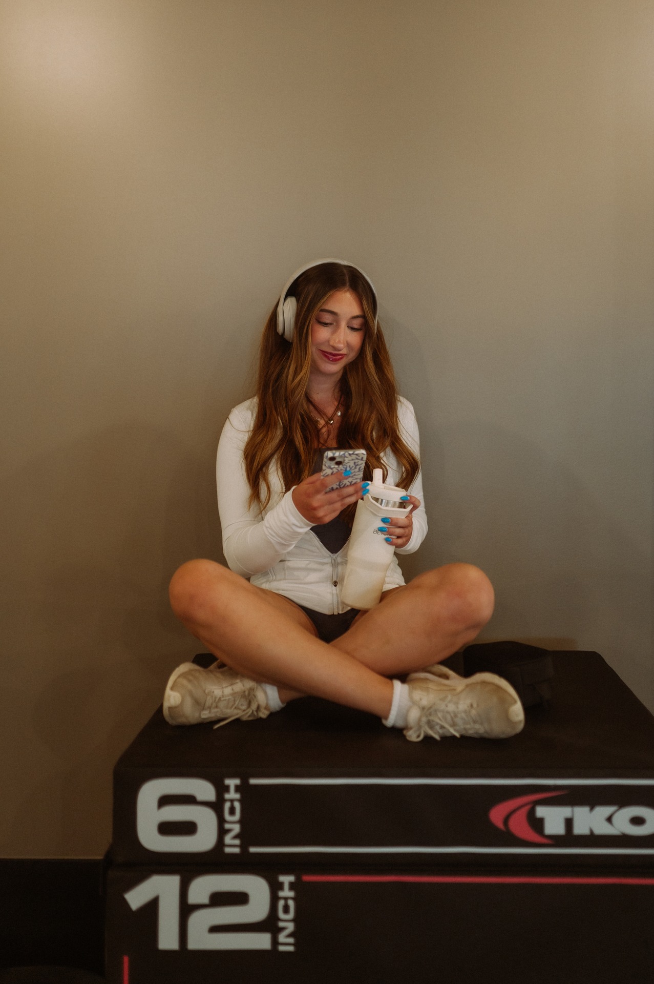 A woman with long hair, wearing headphones, a white top, shorts, and sneakers, sits cross-legged on a gym box in her Purdue off campus housing. She holds a drink in one hand and looks at her phone, smiling. The wall behind her is plain and beige.
