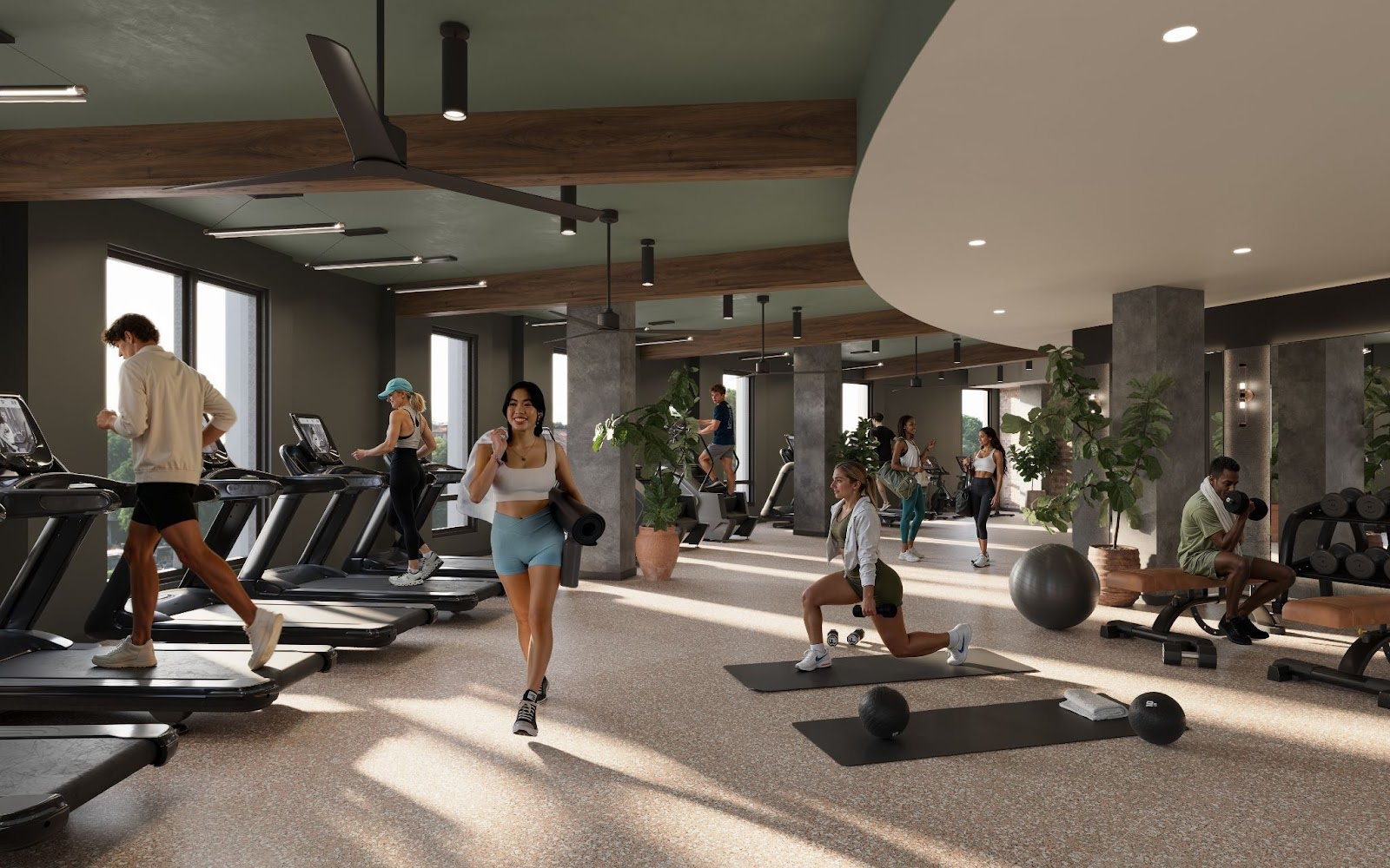 A modern gym at Rambler Riverfront with people working out on treadmills, lifting weights, doing lunges, and various exercises. The space is bright, featuring large windows, plants, and fitness equipment throughout.