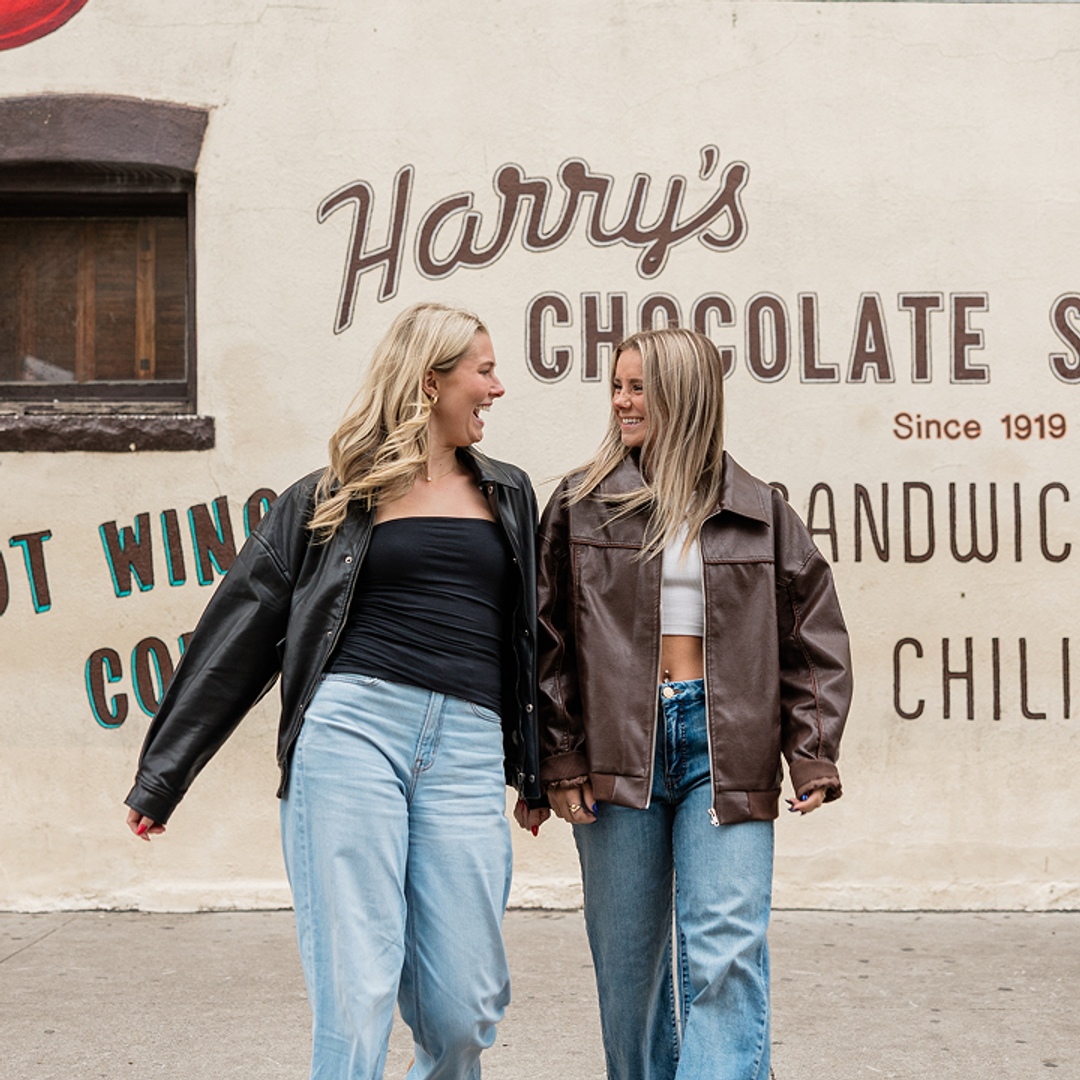 Two women wearing leather jackets and jeans smile at each other while holding hands, walking past the Harry's Chocolate Shop mural near popular apartments in West Lafayette.
