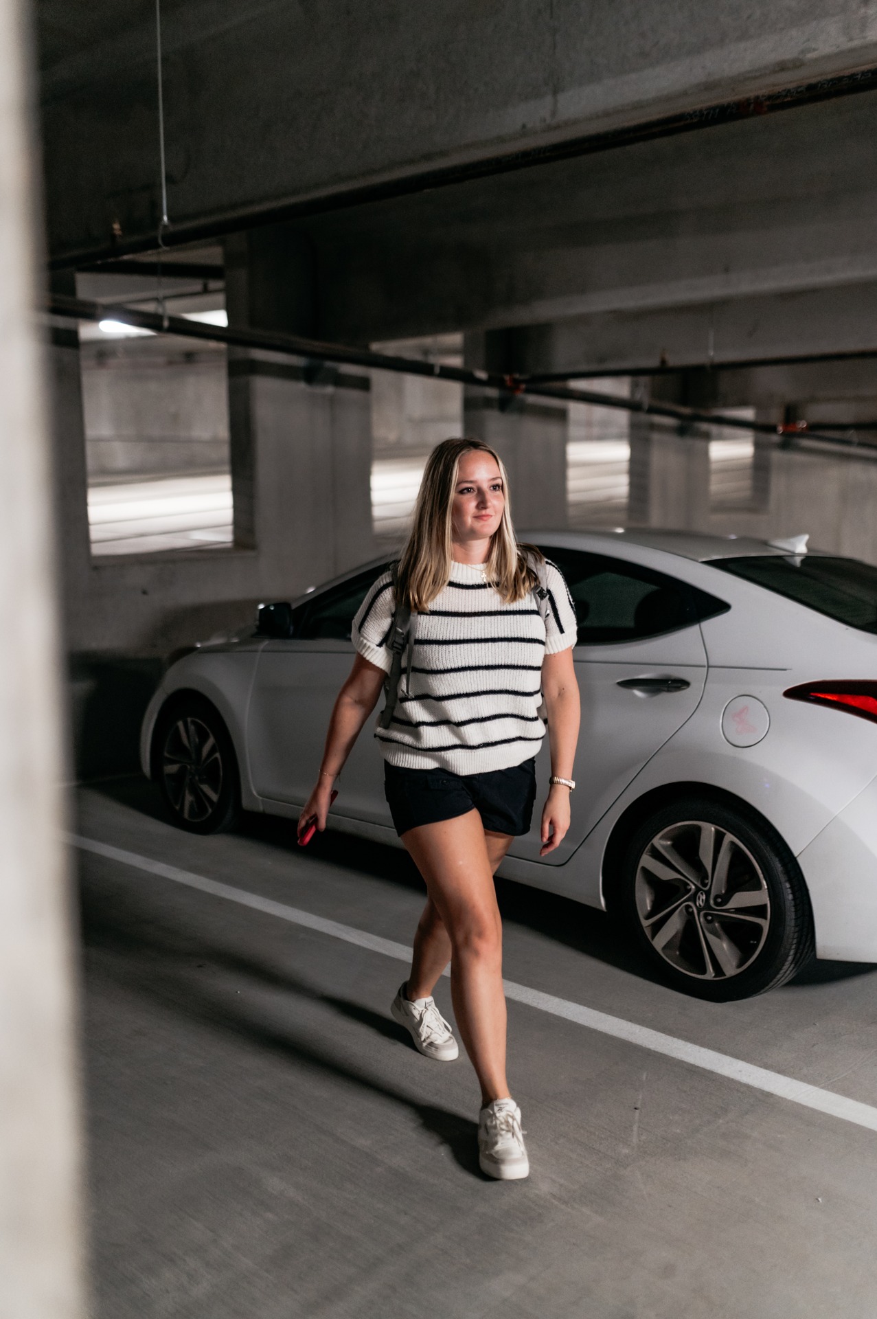 A woman wearing a striped shirt, black shorts, and white sneakers walks in a parking garage next to a white car, holding a pink phone and carrying a backpack—typical of students heading to Purdue off campus housing.