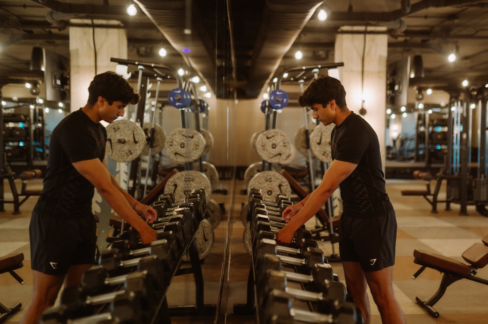 A man in black workout clothes selects a dumbbell from a rack in a gym, his reflection visible in the wall mirror behind him—a modern fitness center often found in Purdue off campus housing. Exercise equipment fills the background.