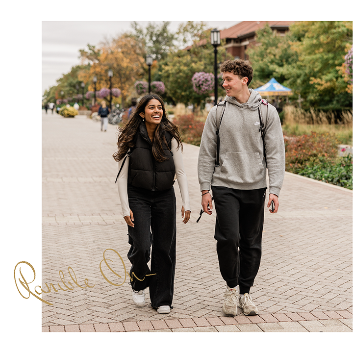 Two young adults walk side by side on a paved campus pathway near Purdue off campus housing, smiling at each other. Both wear casual clothes; the background features trees, flowers, and buildings. A gold cursive signature appears in the lower left corner.