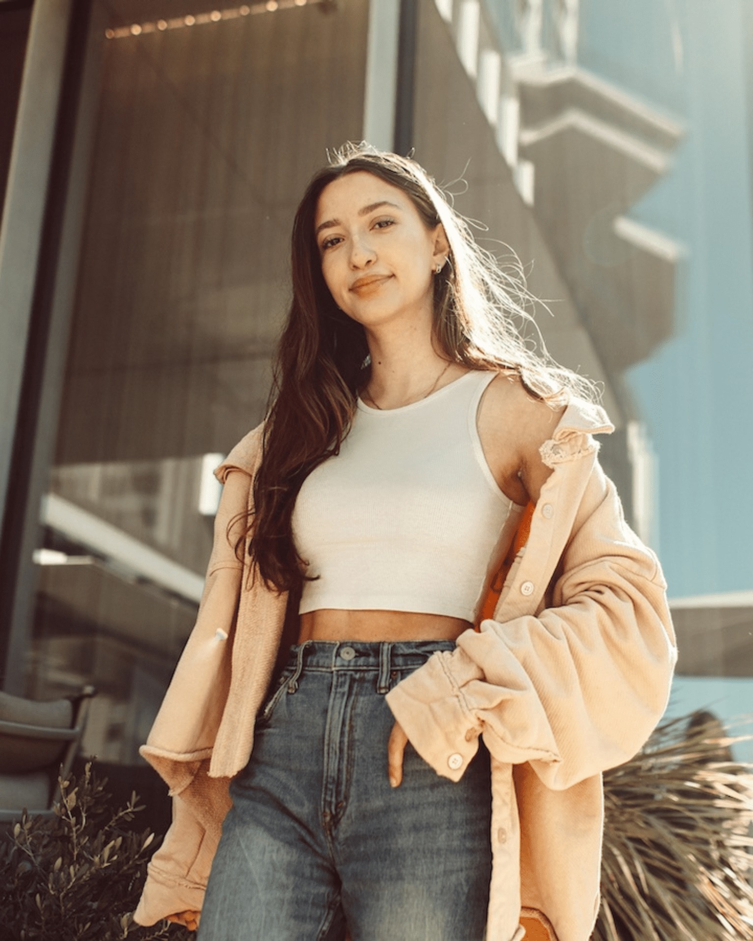 A young woman with long brown hair stands outdoors, wearing a white crop top, high-waisted jeans, and an oversized beige jacket, smiling confidently in front of a modern glass building.