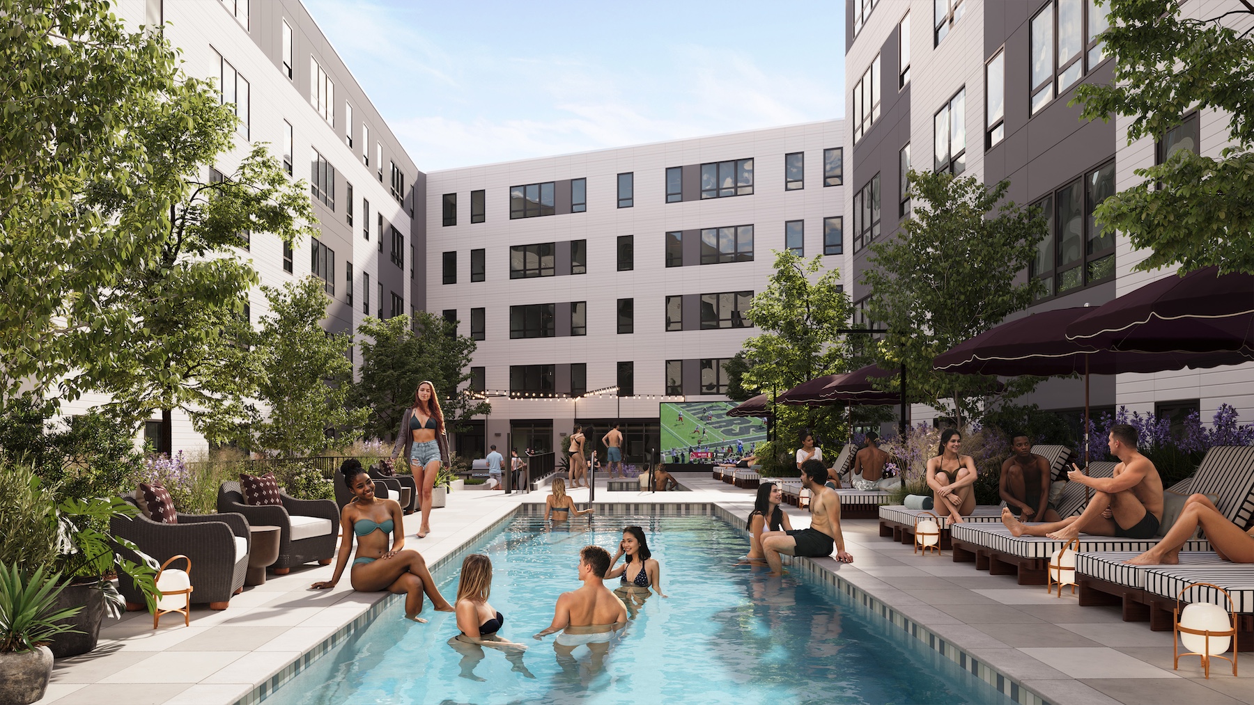 People relax and socialize by an outdoor pool surrounded by lounge chairs and umbrellas in a modern Purdue off campus housing courtyard. Some swim while others gather poolside, enjoying a sunny day among trees and greenery.