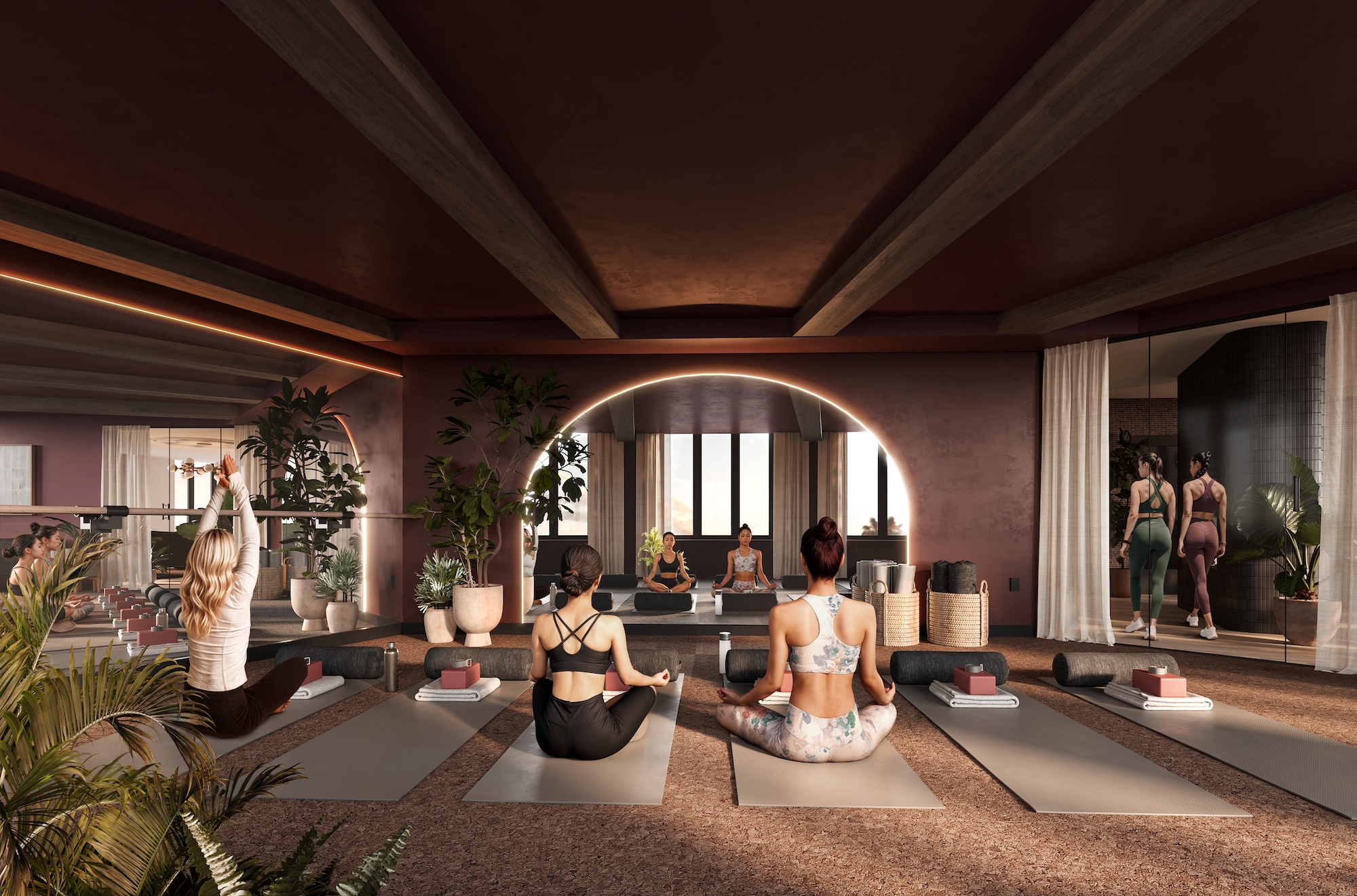 A group of women practice yoga on mats in a stylish, modern studio with plants and soft lighting—perfect for those seeking wellness activities while living in Purdue off campus housing. A mirrored wall reflects participants preparing for the session.