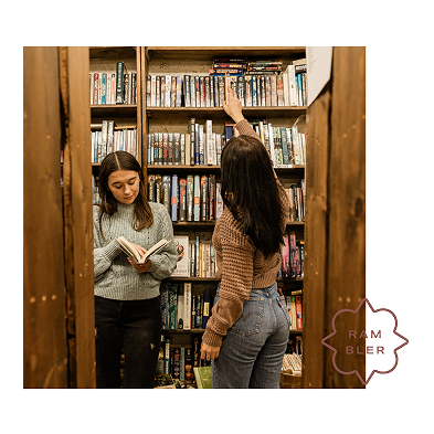 Two women stand between bookshelves in a cozy bookstore, reminiscent of studying at apartments in West Lafayette. One woman reads while the other reaches for a book on the top shelf. Both are dressed in sweaters and jeans, surrounded by shelves brimming with books.