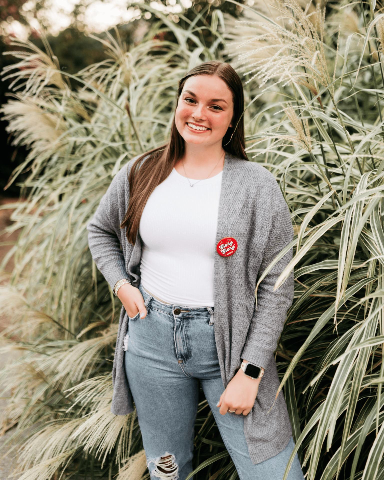 A young woman with long brown hair smiles while standing in front of tall ornamental grass. She wears a gray cardigan, white top, ripped blue jeans, smart watch, and a Happy Birthday button.