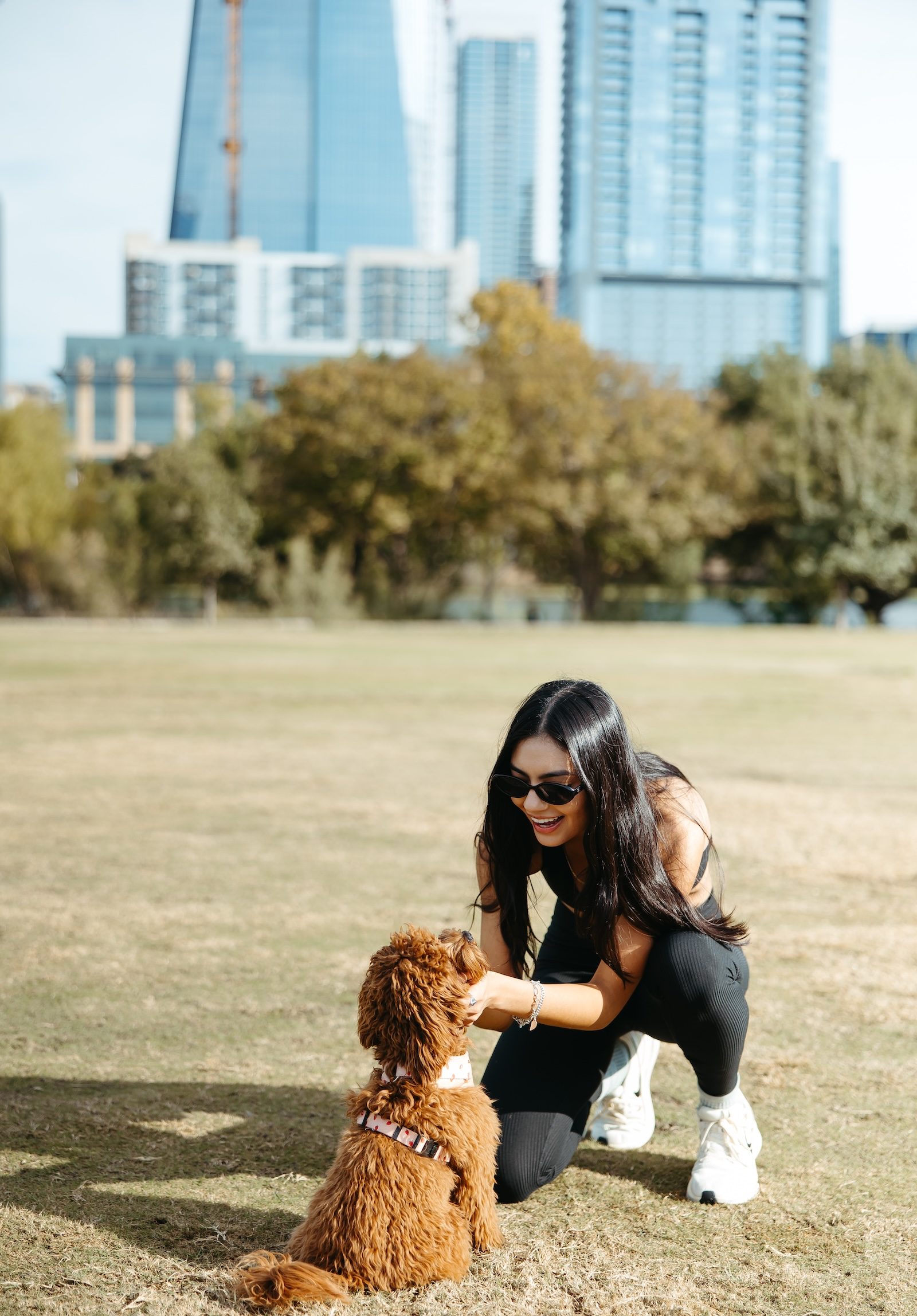 A woman in sunglasses kneels on grass, smiling and petting a small brown dog wearing a harness, with tall buildings and green trees in the background.