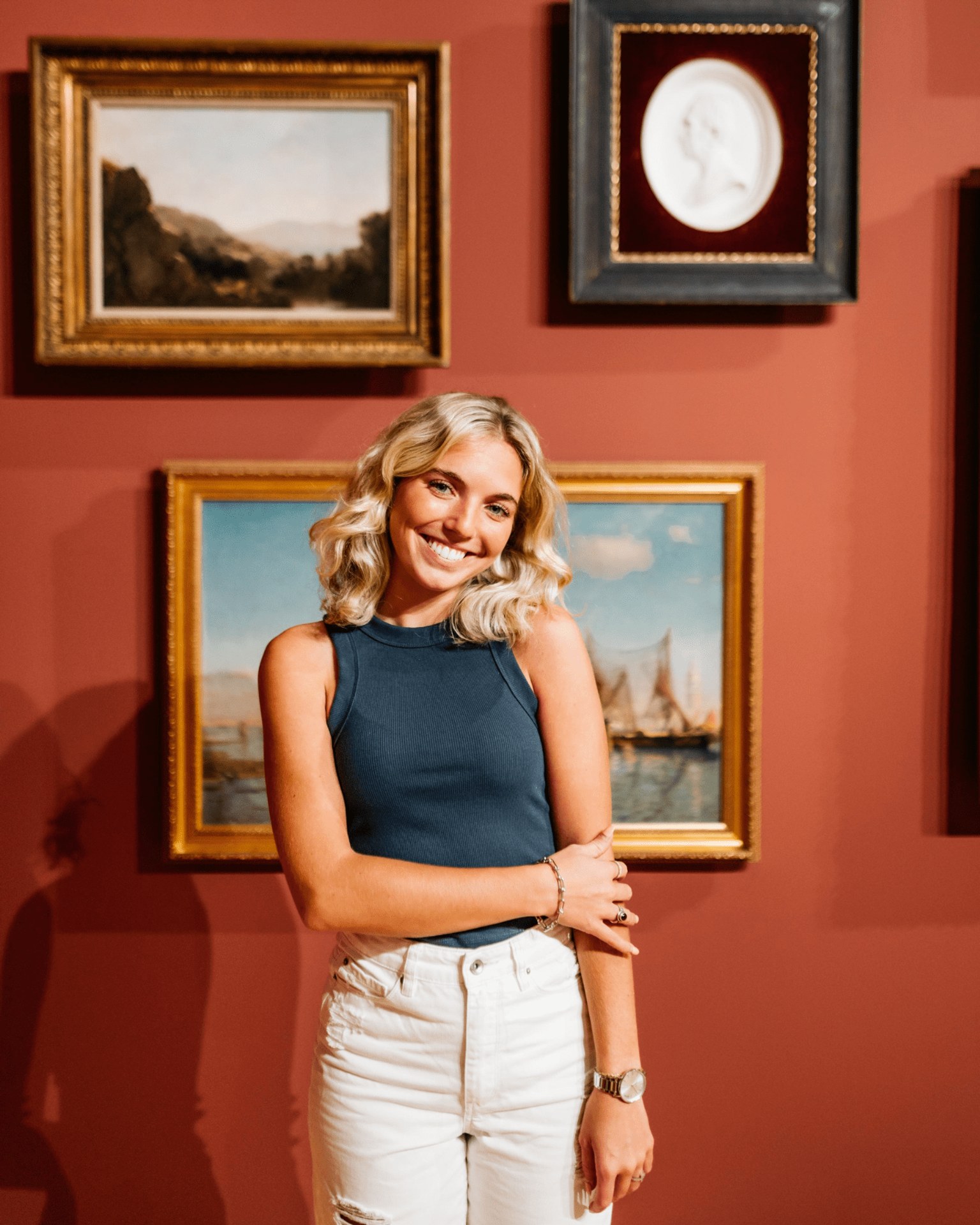 A smiling woman with blonde hair, wearing a sleeveless blue top and white jeans, stands in front of framed artwork on a reddish wall in a gallery.