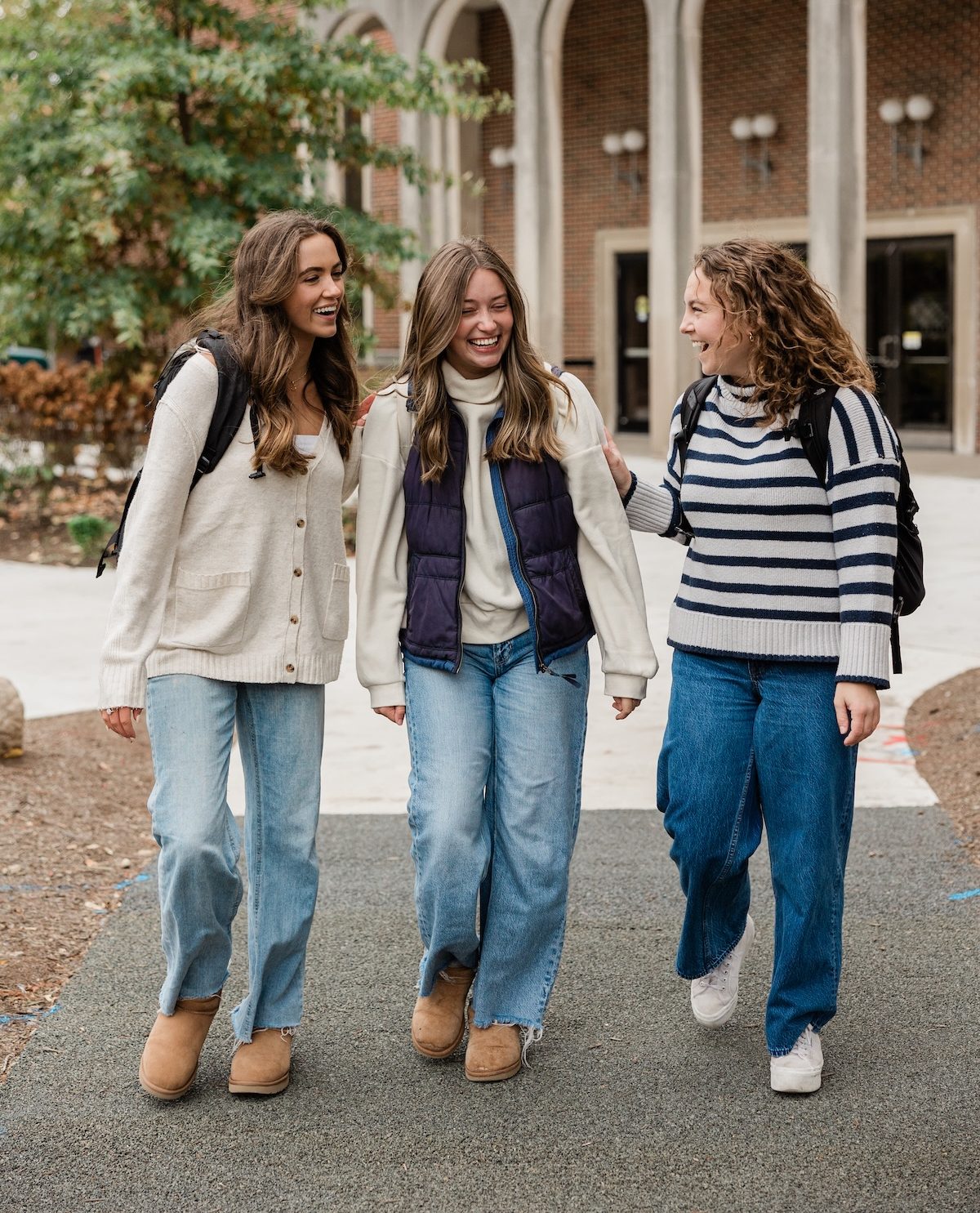 Three young women wearing casual fall outfits and backpacks walk together outside a building, smiling and talking.