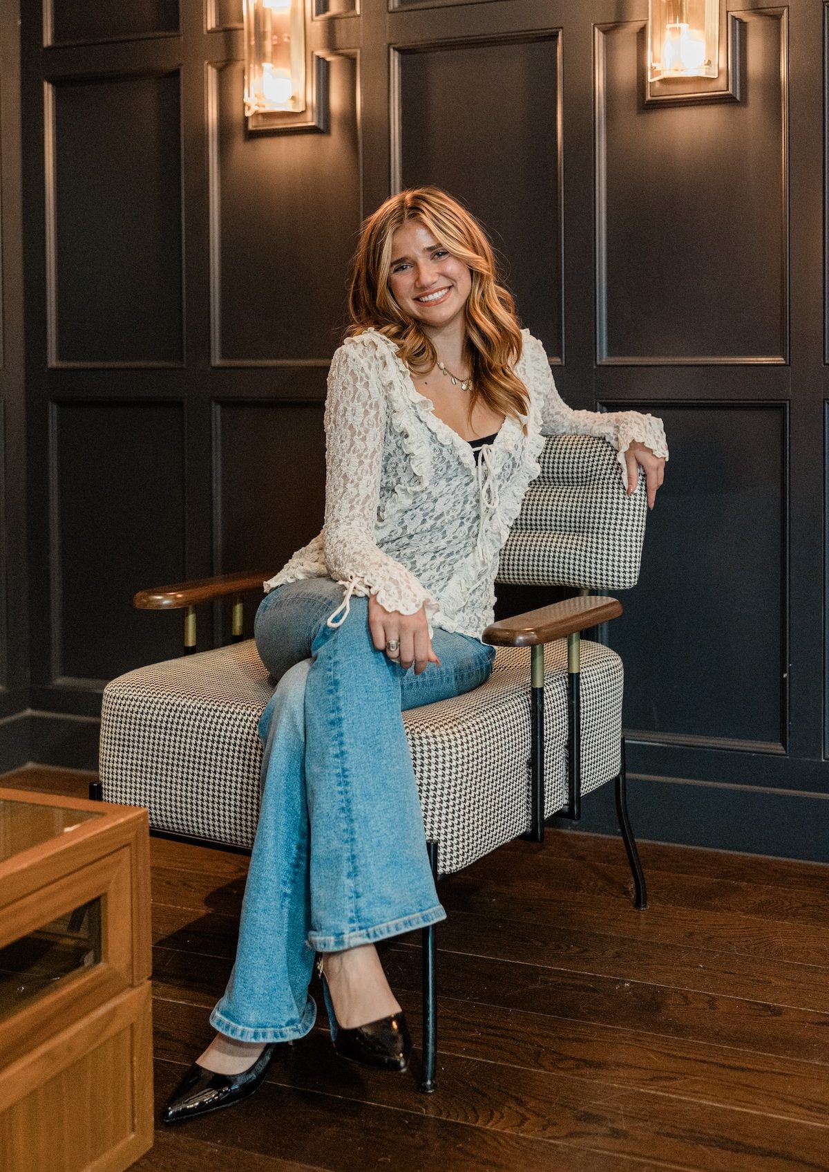 A woman with wavy hair, wearing a white lace top, blue jeans, and black heels, sits comfortably on a patterned armchair. She smiles and poses in a room with dark paneled walls and wooden floors.