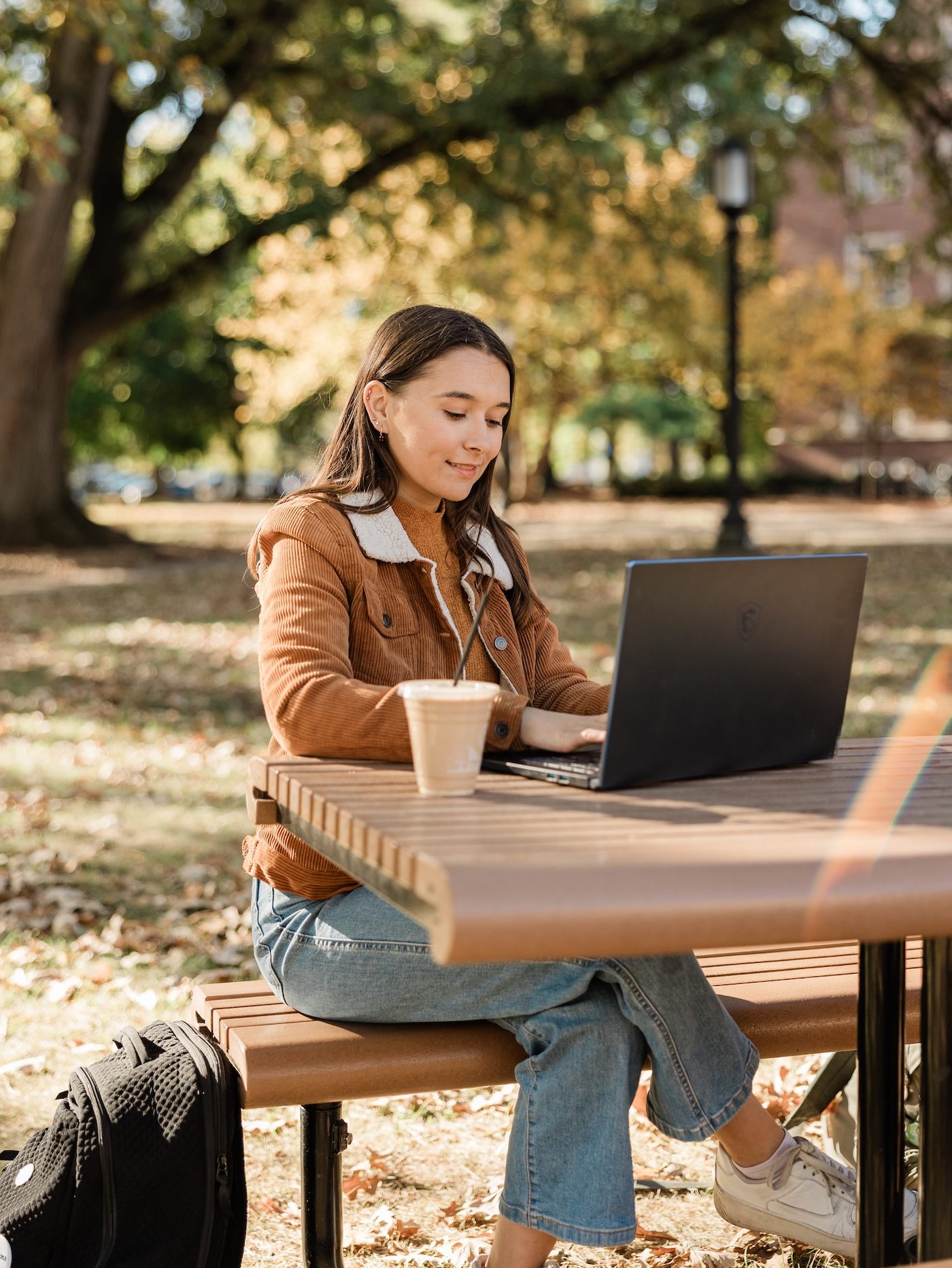 A woman sits at a wooden picnic table in a park, working on a laptop. She has a drink beside her, and a backpack on the ground. The trees around her have green and yellow leaves.