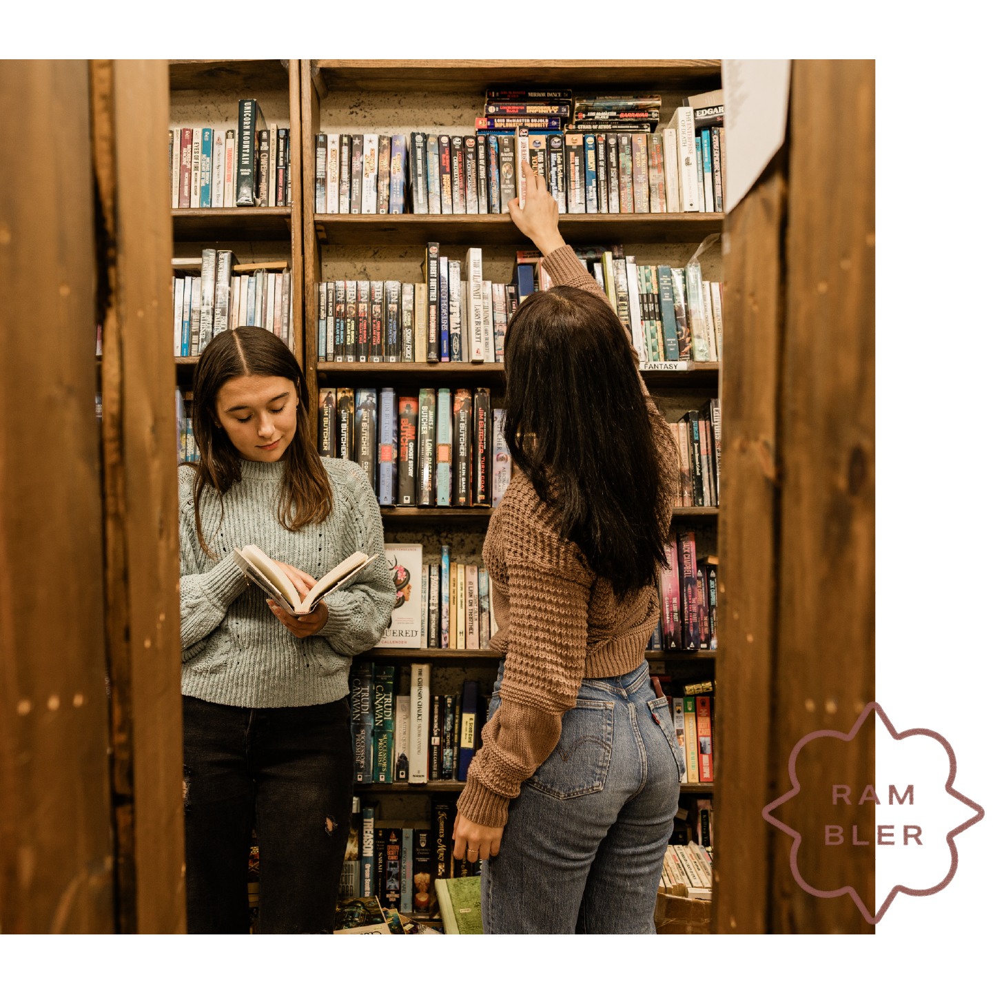 Two women in sweaters stand in a cozy bookstore, reminiscent of the inviting charm found in apartments in West Lafayette. One reads a book while the other reaches for a high shelf, surrounded by shelves of colorful books. A RAMBLER logo is visible in the corner.
