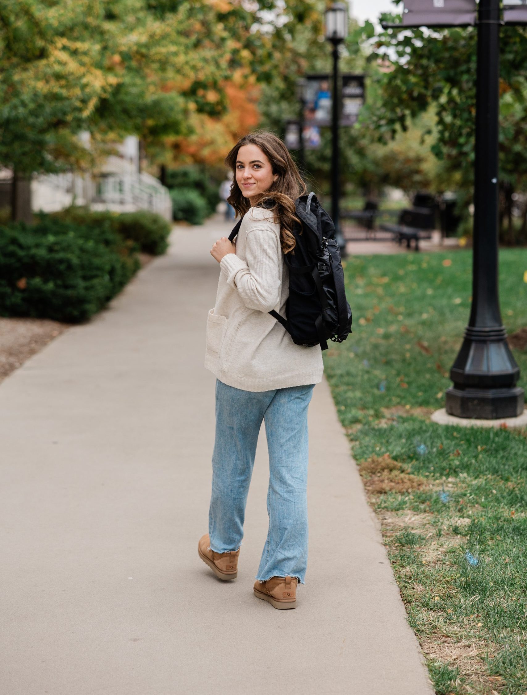 A young woman with long hair, wearing a beige sweater, light blue jeans, and brown shoes, walks on a sidewalk through a park while looking back and smiling. She carries a black backpack.
