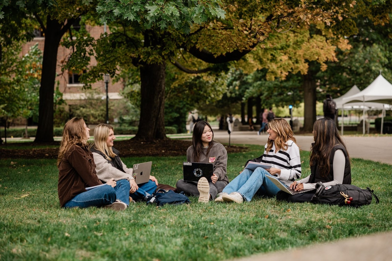 Five young women sit in a circle on the grass, talking and studying with laptops and notebooks. They are outdoors under large trees, with buildings and a tent visible in the background.