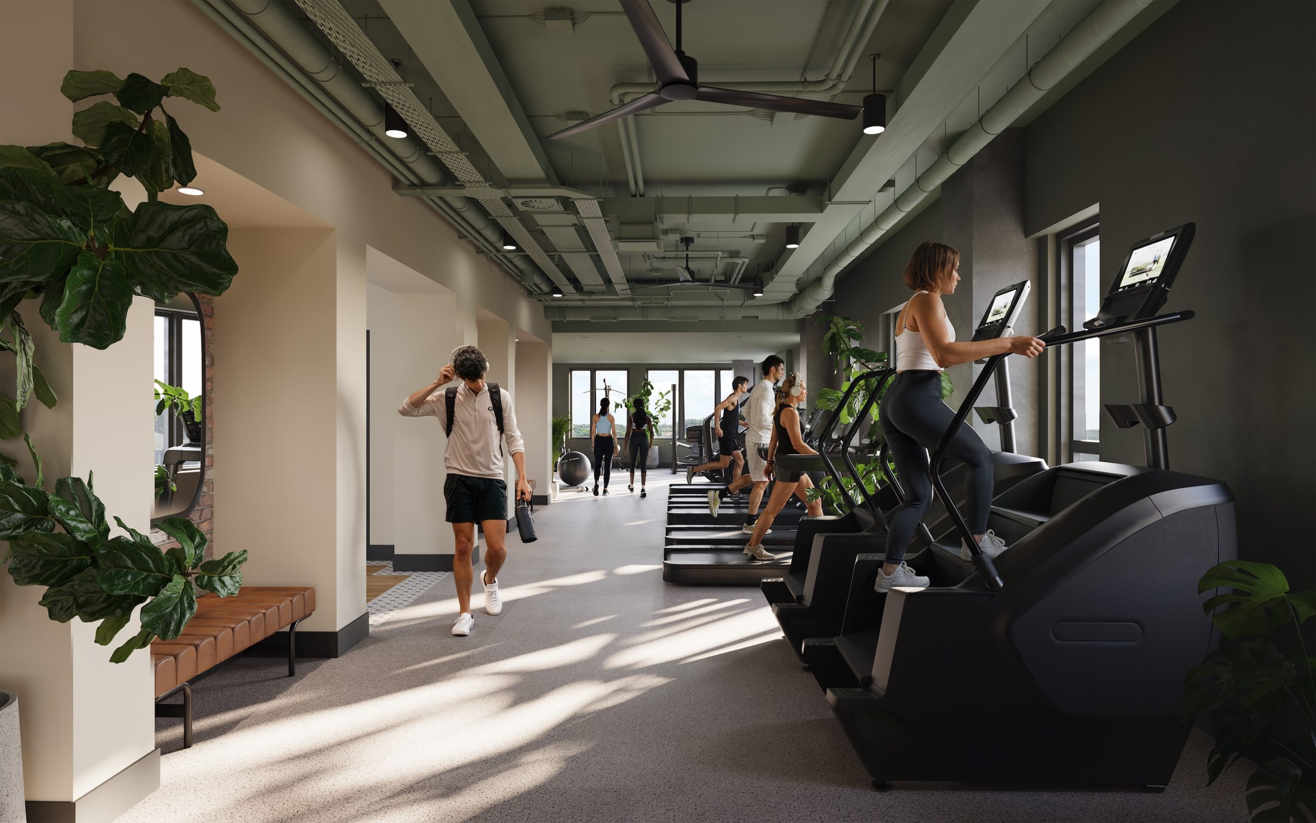 People exercising on stair machines and treadmills in the modern Rambler Riverfront gym, featuring large windows, plants, light walls, and plenty of natural light. Some individuals are walking, while others use the equipment.