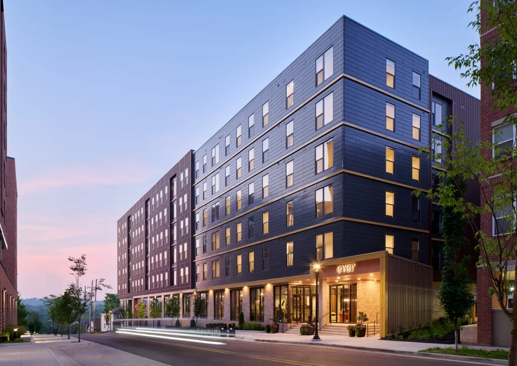 Modern multi-story Chauncey Square apartments with large windows and a ground floor entrance, illuminated at dusk. The street in front features blurred lights from passing vehicles, while trees line the sidewalk.