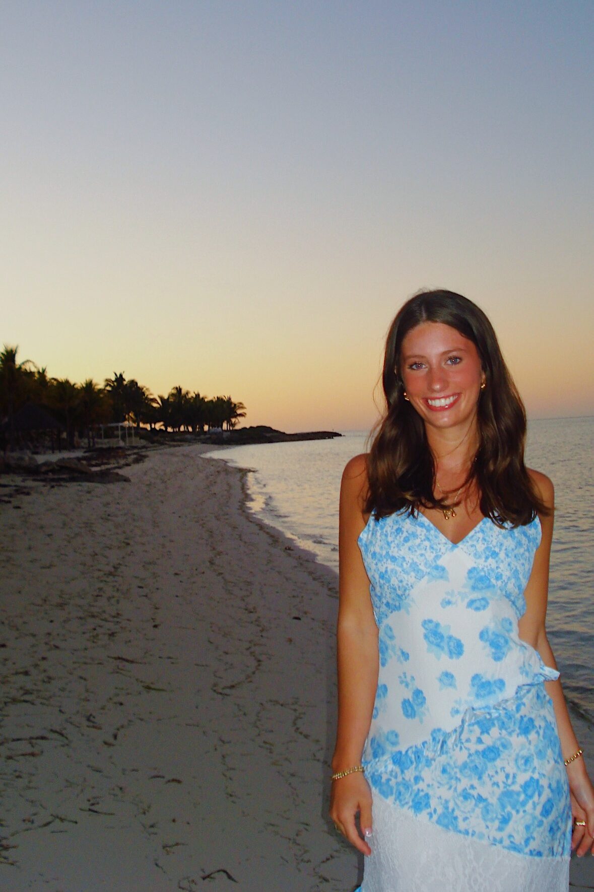 A woman in a white and blue floral dress smiles while standing on a sandy beach at sunset, dreaming of student apartment jobs in West Lafayette, with calm water and palm trees in the background.
