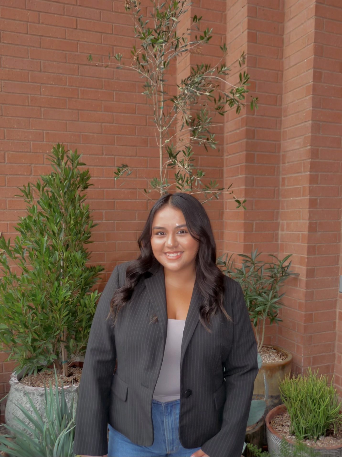 A woman with long, wavy dark hair, wearing a black pinstripe blazer and jeans, stands smiling in front of green potted plants and a brick wall—perfect inspiration for student apartment jobs in West Lafayette.