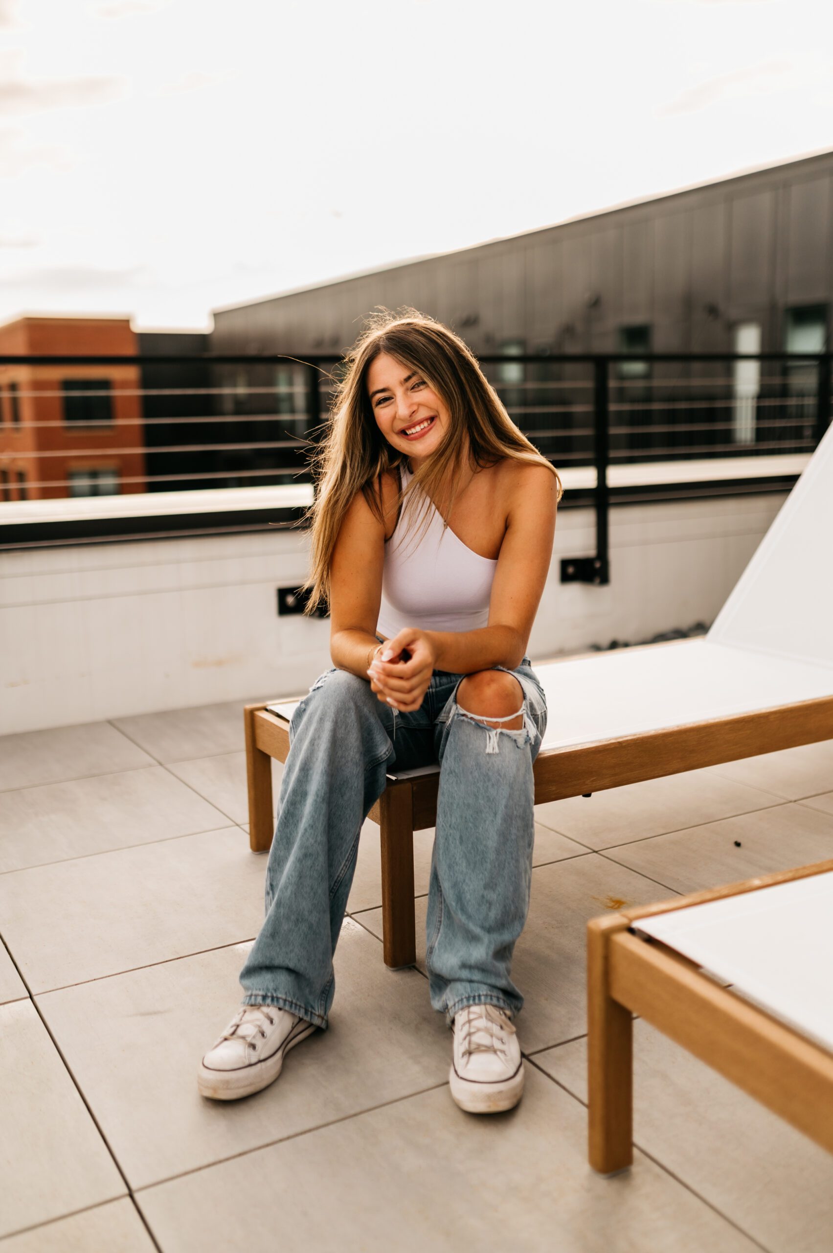 A young woman with long hair sits on a lounge chair outdoors, smiling at the camera. On a modern rooftop patio, she looks relaxed in her white one-shoulder top and ripped jeans—perhaps enjoying a break from student apartment jobs in West Lafayette.