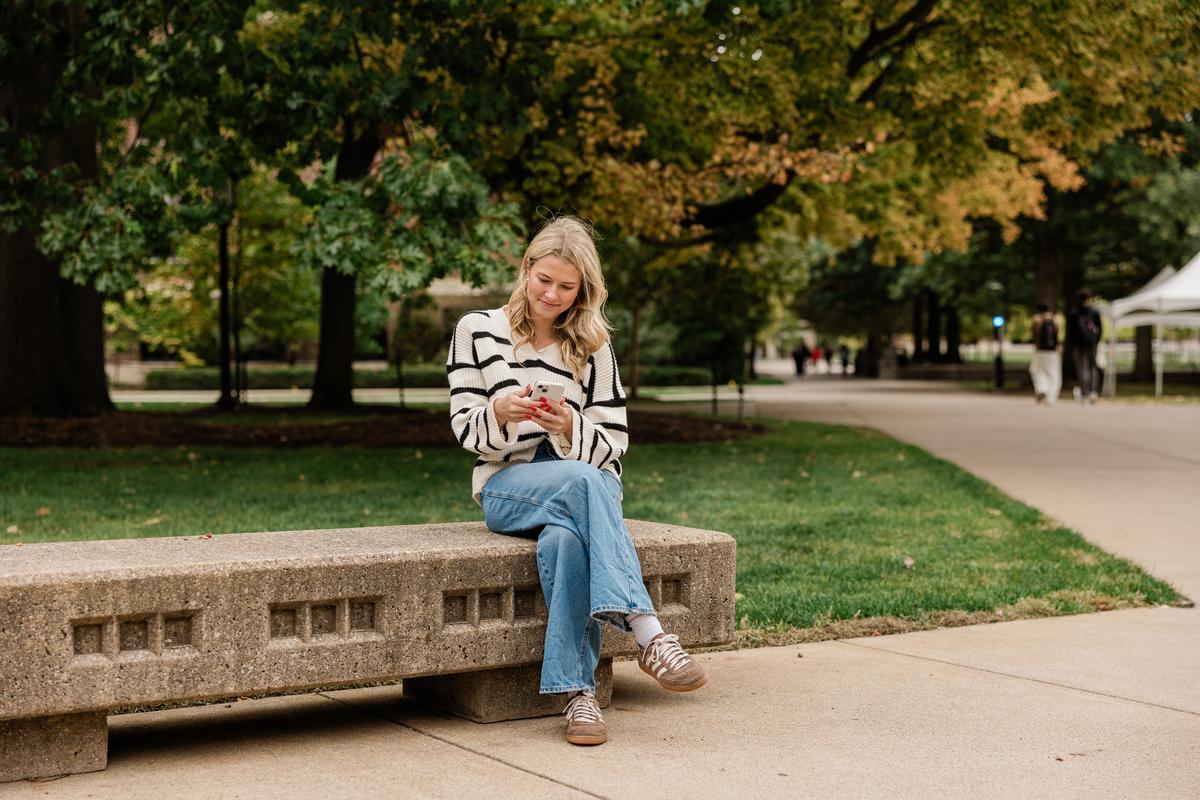 A young woman in a striped sweater and blue jeans sits on a concrete bench in a park, smiling at her phone—perhaps reading about the Rambler Riverfront vs Ever debate. Trees and a walking path are visible in the background.