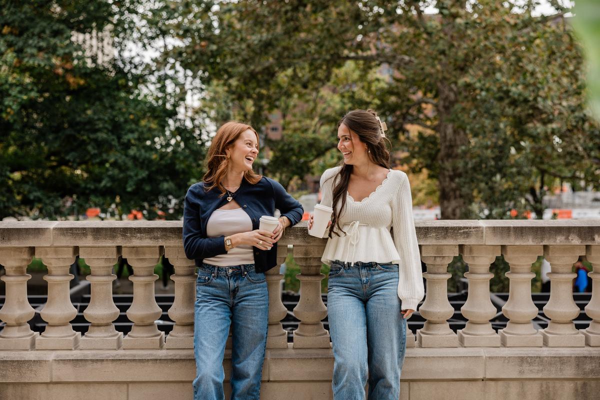 Two women stand and chat, smiling at each other with coffee cups in hand, casually dressed by a stone balustrade. Their relaxed vibe makes for a scene reminiscent of a friendly debate: rambler riverfront vs verve. Lush greenery fills the background.