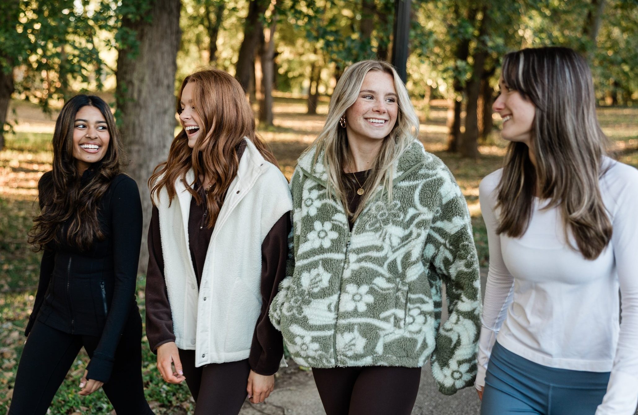 Four young women walk together outdoors on a tree-lined path in West Lafayette, smiling and talking about student apartment jobs as they enjoy a pleasant day in the park, dressed in casual, warm clothing.