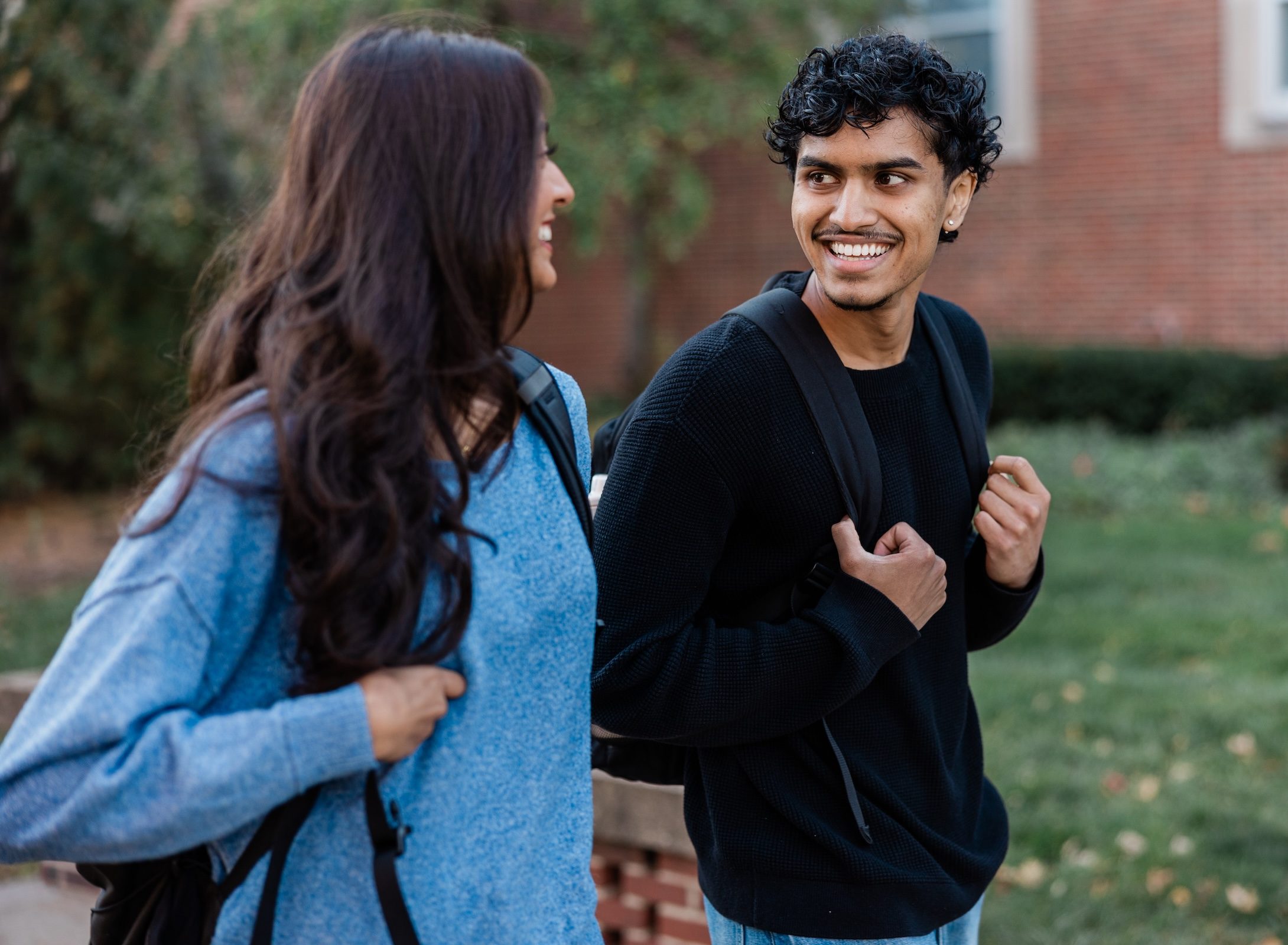 Two students with backpacks walk outdoors, smiling at each other on a campus with greenery and a brick building in the background, perhaps discussing student apartment jobs in West Lafayette.