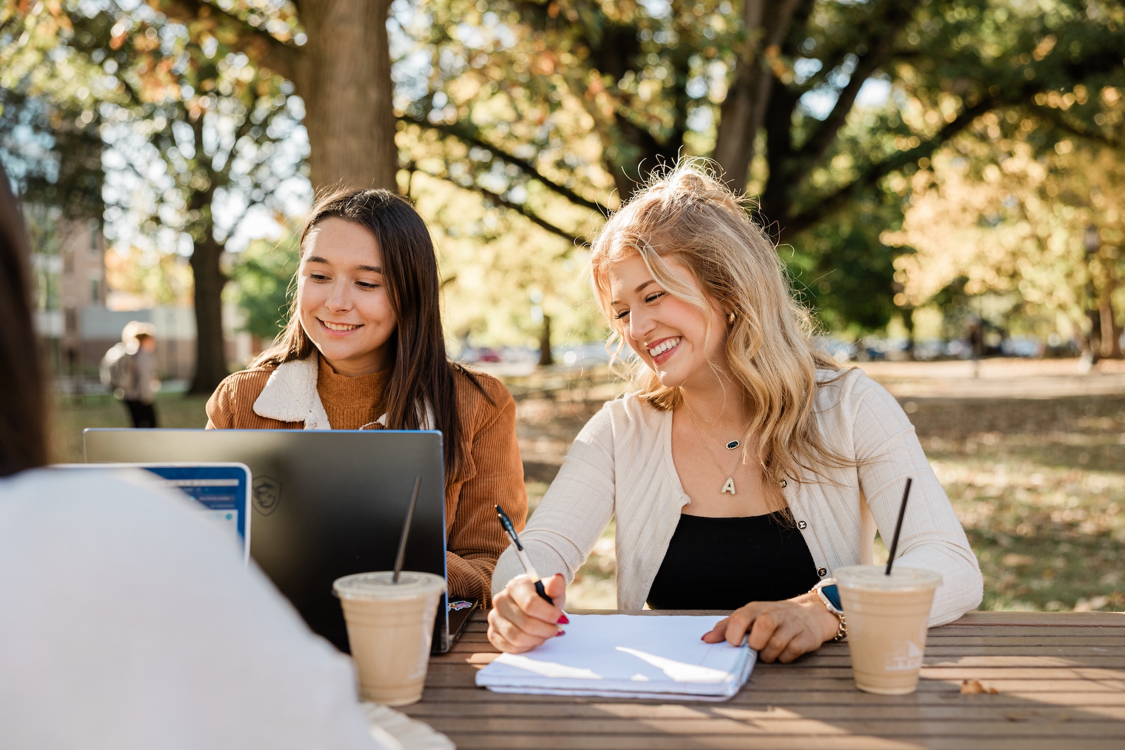 Two young women sit at a table outside their student apartment in West Lafayette, smiling as they work together; one types on a laptop while the other writes on paper. Two iced coffee drinks are on the table, with trees and sunlight in the background.