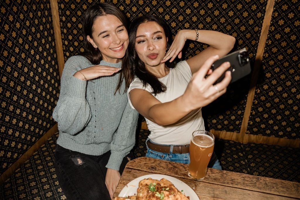 Two girls sit in a restaurant booth, taking a selfie together with food and drinks on the table.