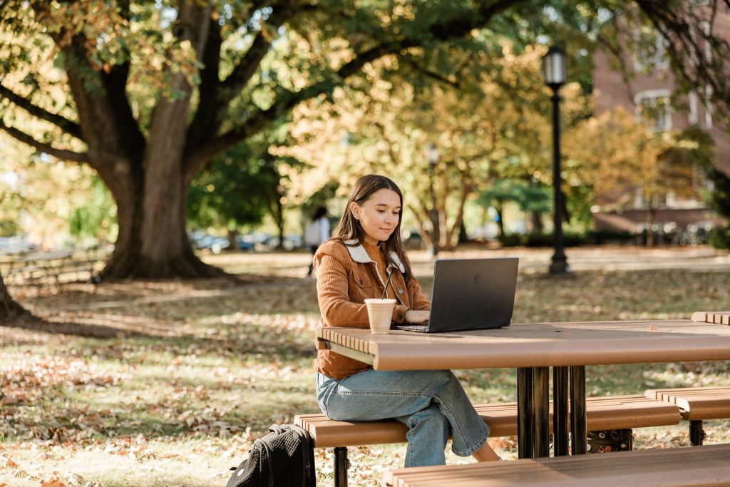 A college girl sitting at a picnic table outdoors, working on her laptop.