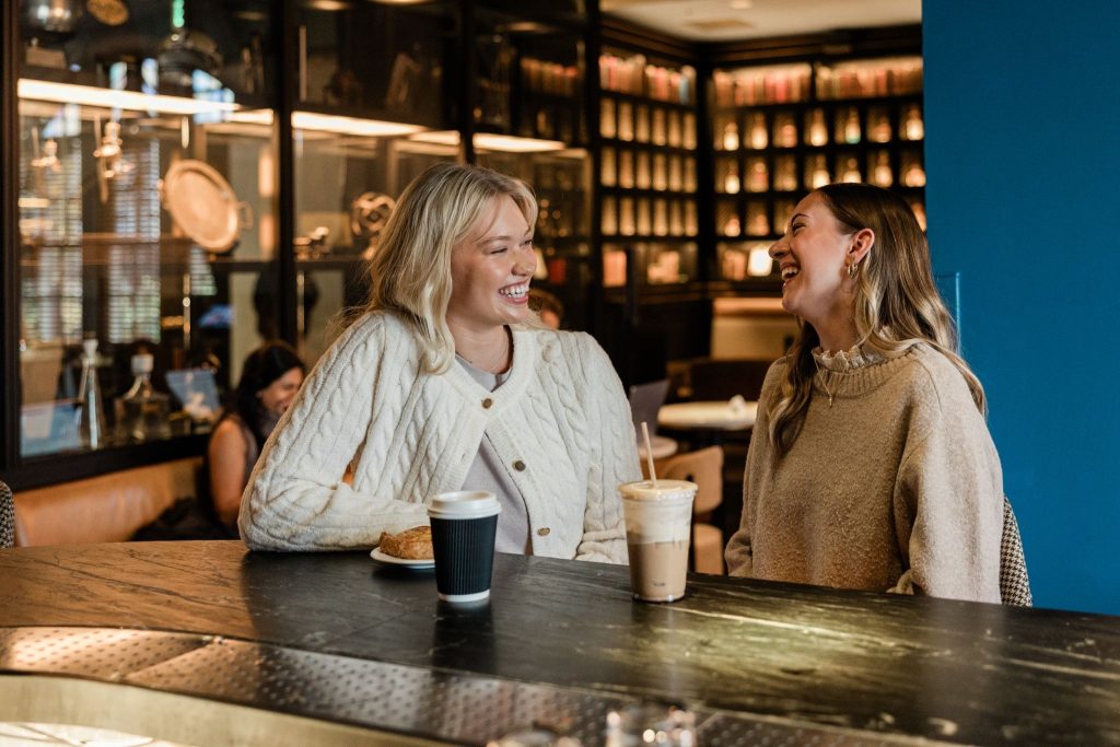 Two college girls sitting at a coffee shop, talking and smiling together over drinks.