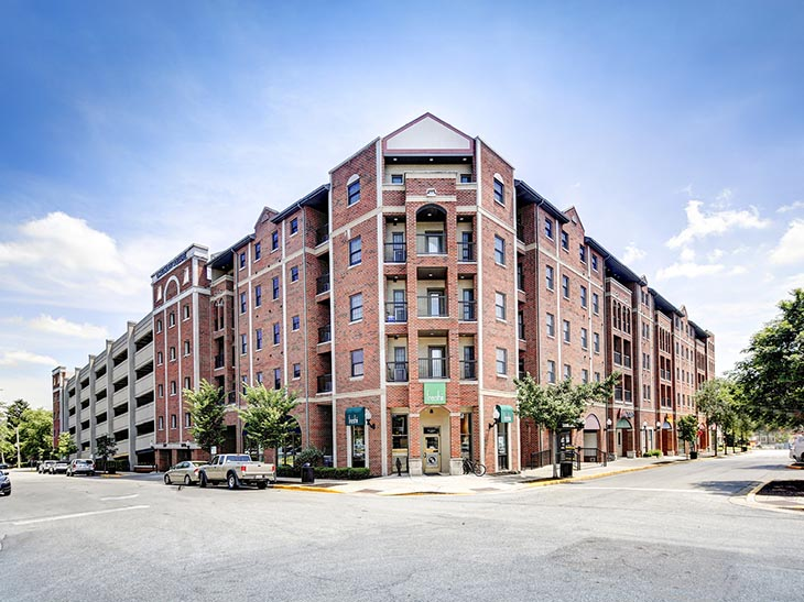 A large, multi-story red brick apartment building with balconies sits on a street corner under a blue sky, offering a vibe reminiscent of Rambler Riverfront vs Chauncey Square. Parked cars and trees line the sidewalks.