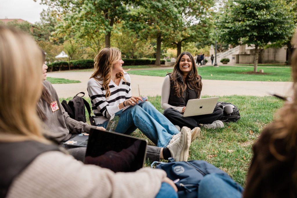 A group of Purdue Students sitting on the grass by the engineering fountain, chatting while using their laptops together.