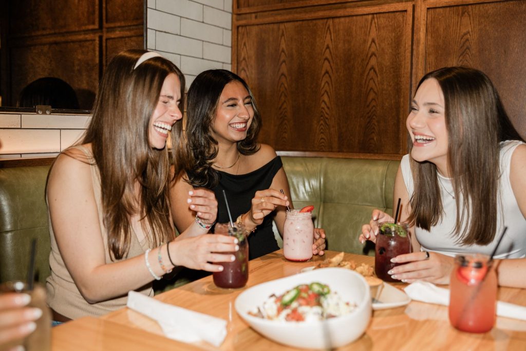 A group of friends sitting at a restaurant table with food and drinks, laughing and enjoying each other’s company.