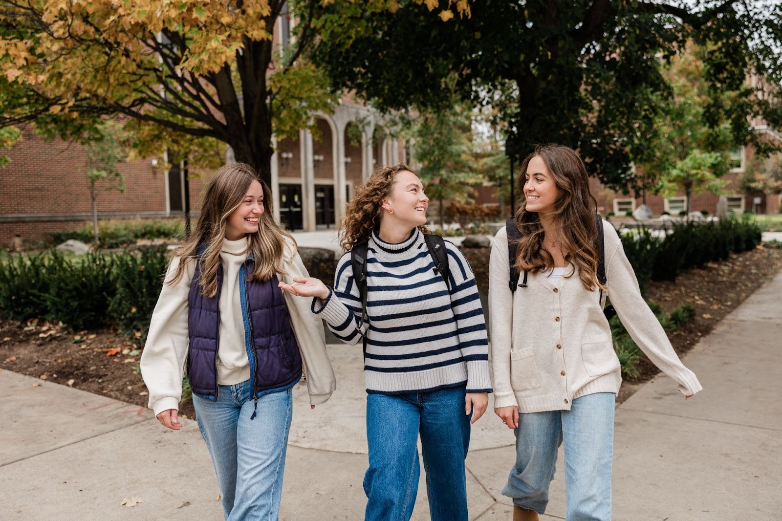 Three young women wearing casual fall outfits and backpacks walk together on a college campus sidewalk, smiling and talking about roommate matching at Purdue, with trees and a brick building in the background.