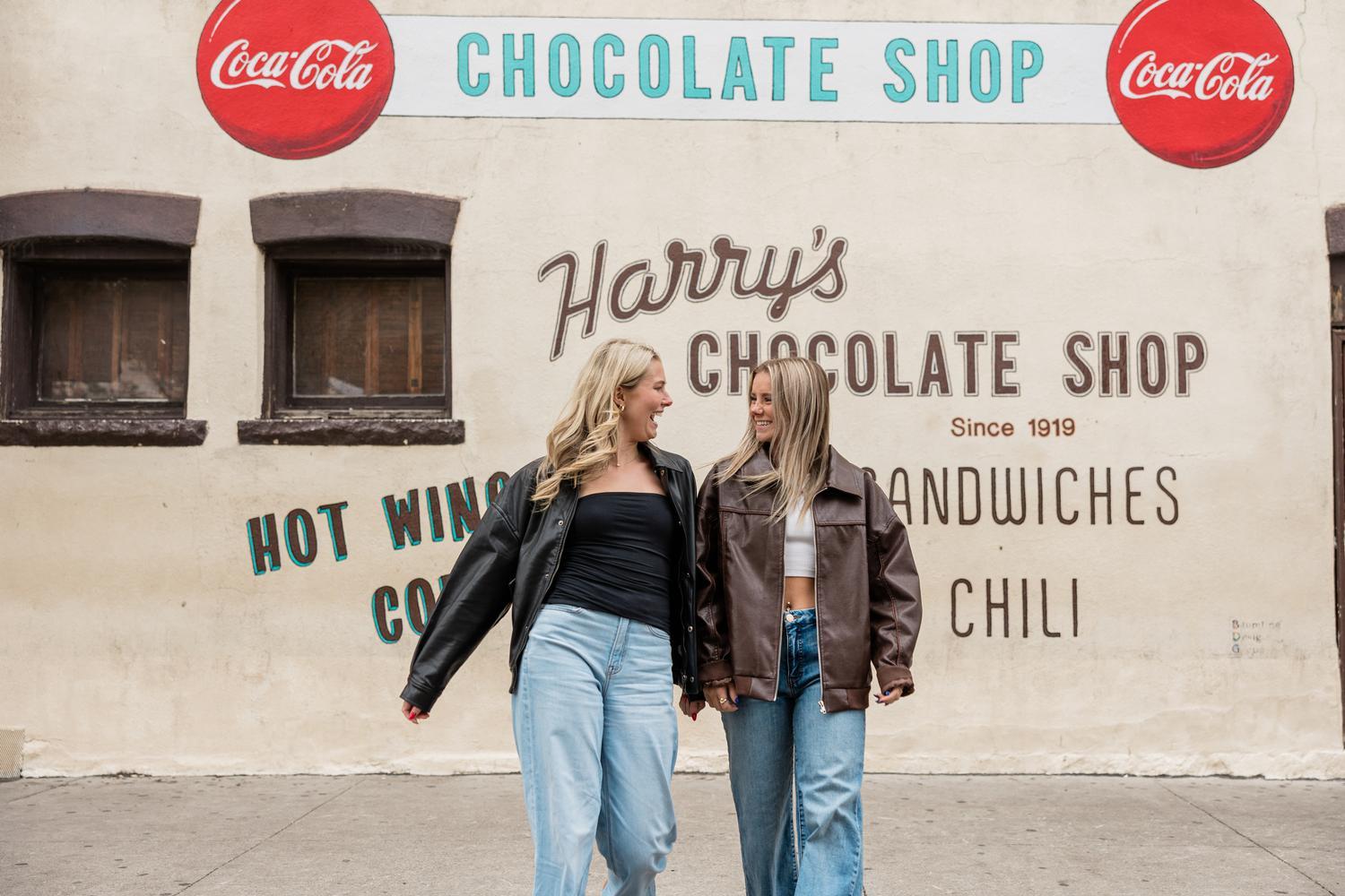 Two young women, holding hands and smiling in front of a vintage Harry's Chocolate Shop sign with Coca-Cola logos, share a moment after reading an Ever West Lafayette apartment review. Both wear casual clothes and leather jackets.