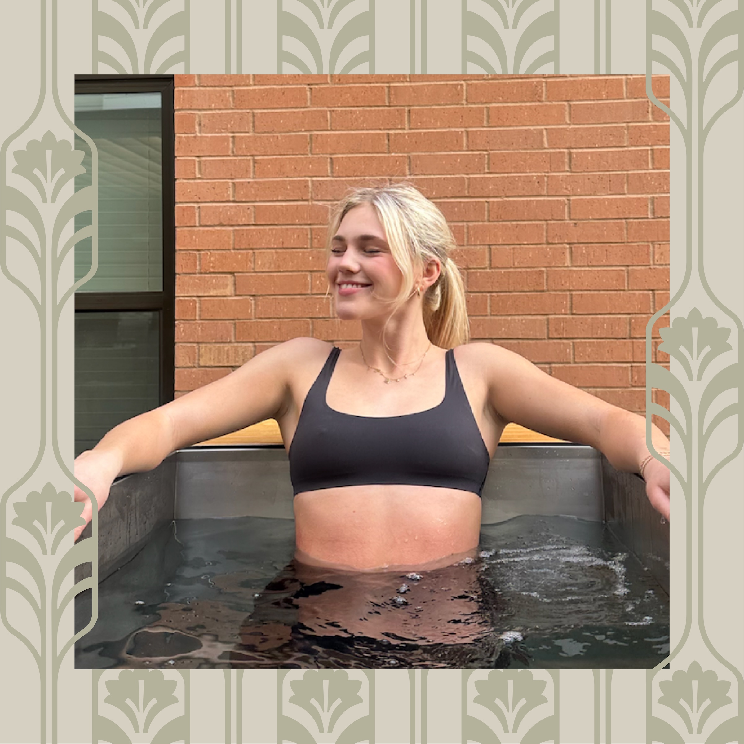 A smiling woman with blonde hair in a ponytail relaxes in a black sports bra, sitting in an outdoor ice bath near Purdue off campus housing, brick and window in the background, framed by a decorative patterned border.
