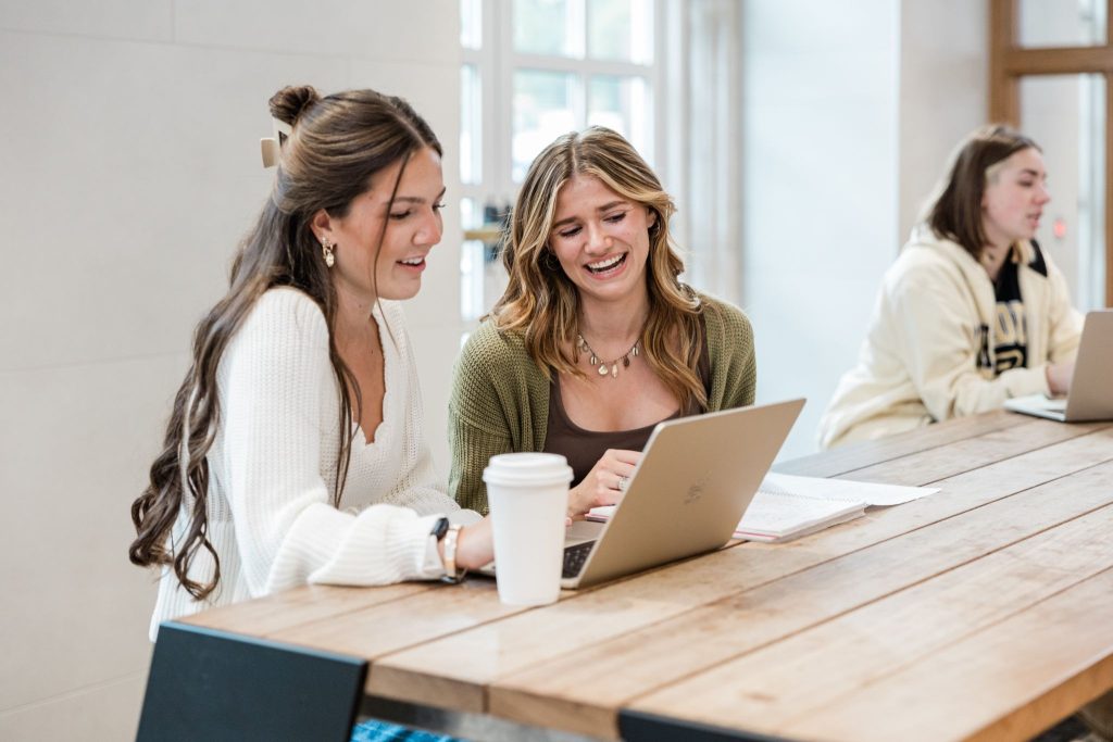 Two women sitting together, looking at a computer screen while holding coffee cups.