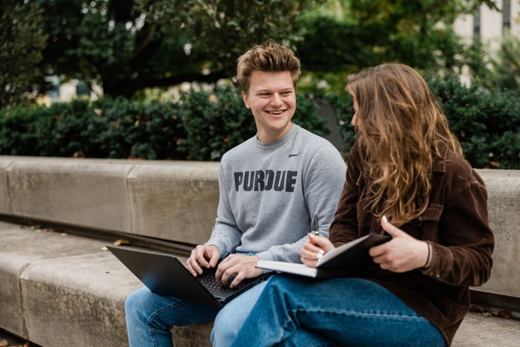 A boy and a girl sit on steps, talking while using their laptops and notebook.