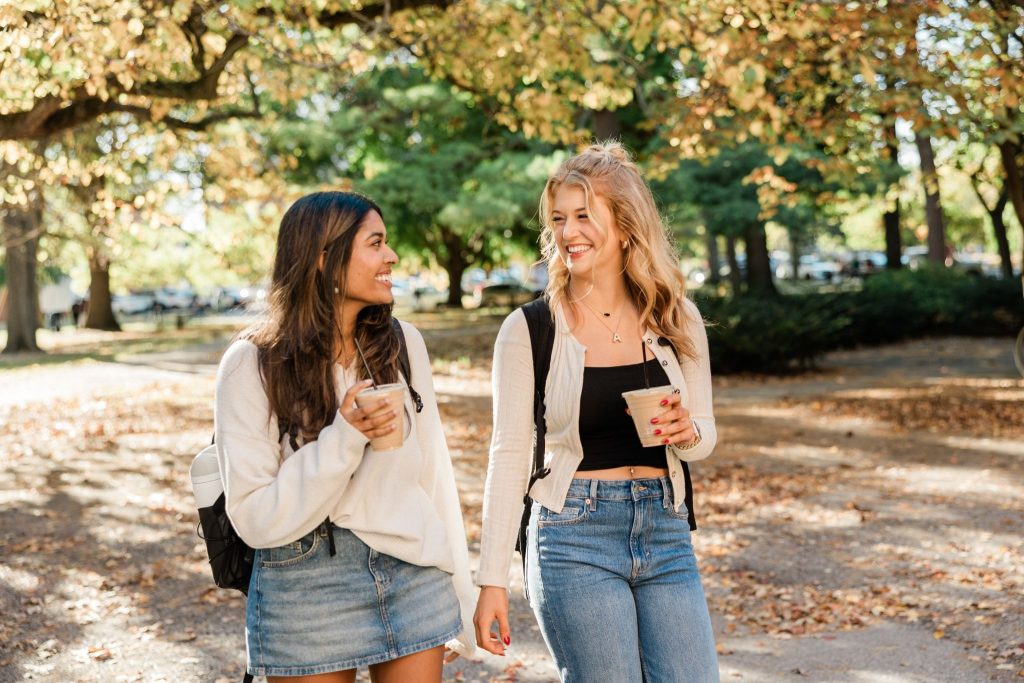 Two girls walk together, wearing backpacks, chatting and holding coffee cups.