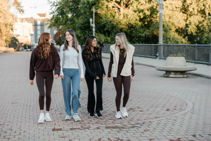 Four young women walk together outside on a paved path near Purdue parking, smiling and talking. Trees and buildings are in the background, and they are dressed casually in jackets, sweaters, and sneakers.