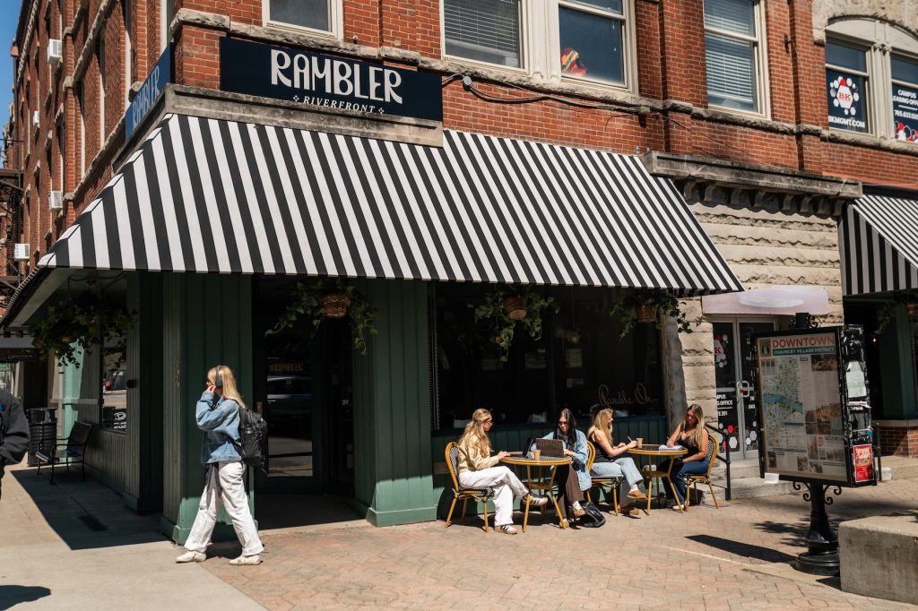 People sit at tables outside a café called Rambler on a sunny day, perhaps pausing during their rambler riverfront tour. One person walks past, while others converse. The café has a striped awning and is nestled on a street corner lined with brick buildings.