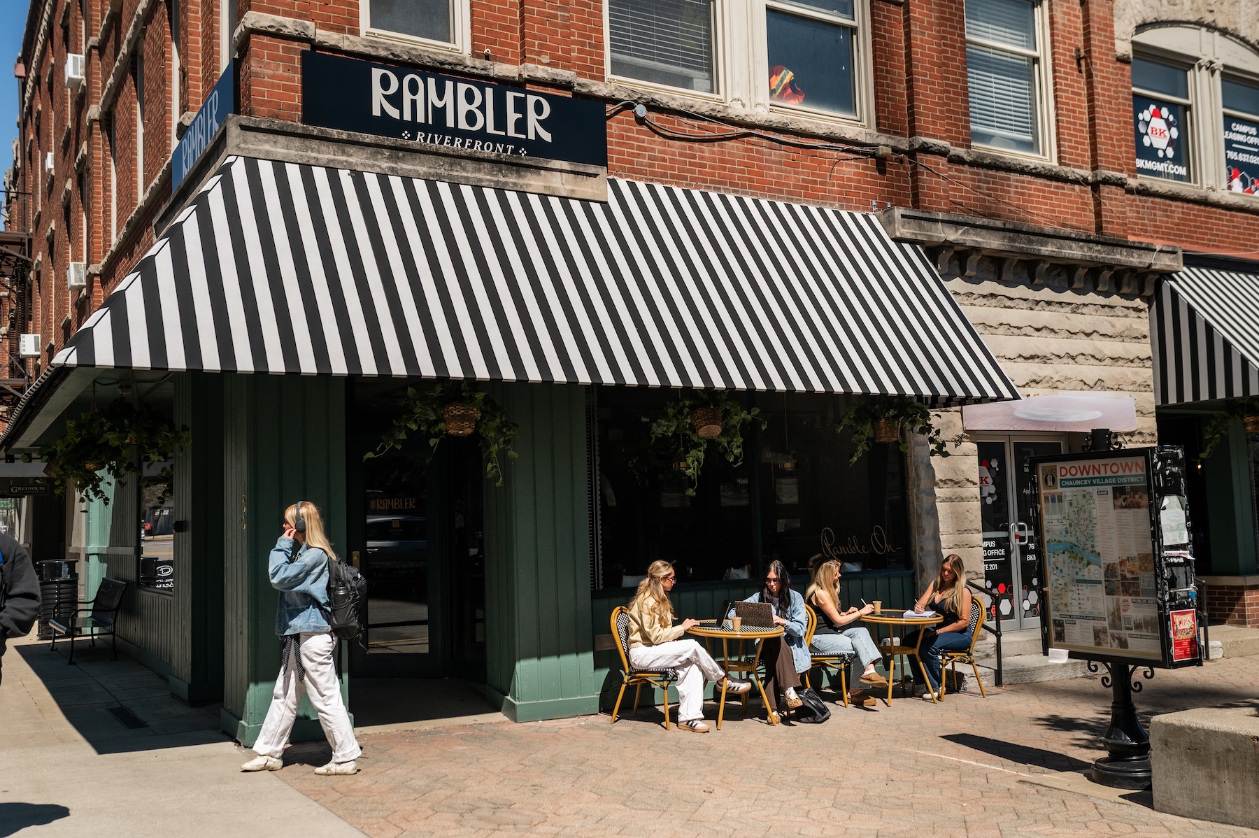 People sit at tables outside a café called Rambler on a sunny day, perhaps pausing during their rambler riverfront tour. One person walks past, while others converse. The café has a striped awning and is nestled on a street corner lined with brick buildings.