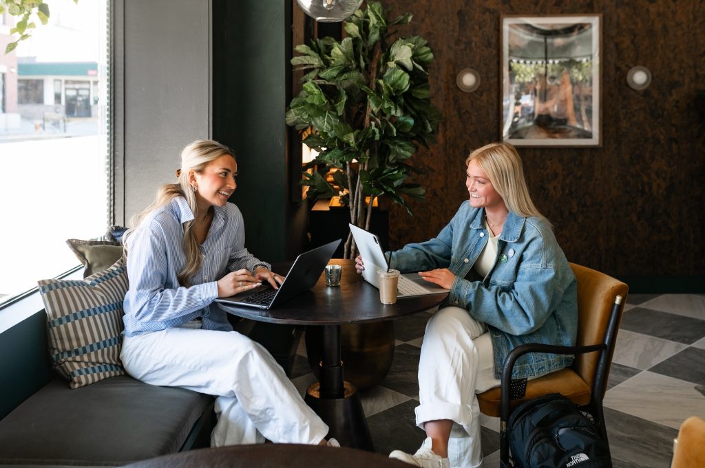 Two women sit at a round table in a modern café, smiling and working on laptops as they plan their rambler riverfront tour. One drinks coffee, and they are surrounded by plants and natural light from a large window.